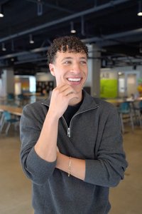 A young man with curly hair smiles and looks to the side while standing with one hand near his mouth in a modern indoor.