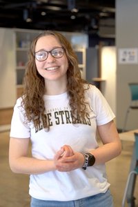 A smiling woman with curly hair and glasses stands indoors wearing a white t-shirt and blue jeans.