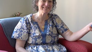 A smiling woman with curly hair sits in a red armchair, looking directly at the viewer with a warm expression.
