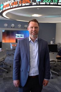 A man in a blue blazer and checkered shirt smiles standing in an office with a circular stock ticker above him.