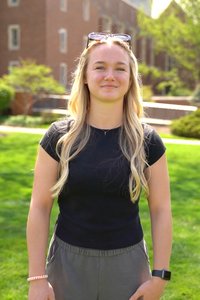 A young woman with long blonde hair stands outside on green grass in front of a brick building, smiling slightly.