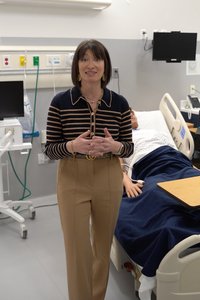 A woman stands in a hospital room speaking with a patient lying in a bed covered by a dark blue blanket.