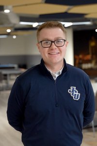 A man wearing glasses and a navy blue zip-up sweater with a logo smiles while standing indoors with ceiling lights above.