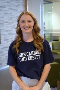 A smiling woman with long curly hair stands indoors wearing a navy shirt that says John Carroll University.