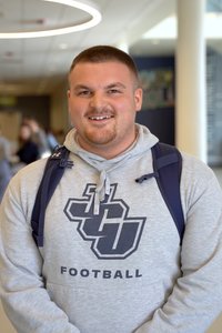 A smiling man wearing a gray hoodie with a football logo and a navy blue backpack stands indoors.