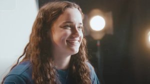 A young woman with long curly hair smiles while looking slightly to the right against a dark background with bright lights.