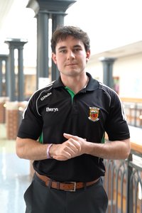 A young man stands indoors with his hands clasped, wearing a black sports shirt with logos and a brown belt.