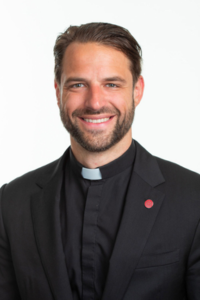 Young priest with brown hair, short beard, and a black suit with a white collar