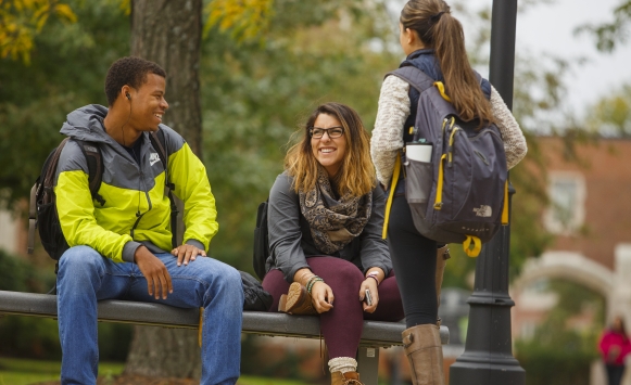 One male and two female students talking on a bench near the quad in the fall